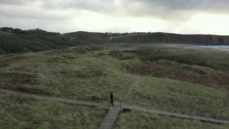 man walking on boardwalk by fields towards playa de xago beach in asturias, spain