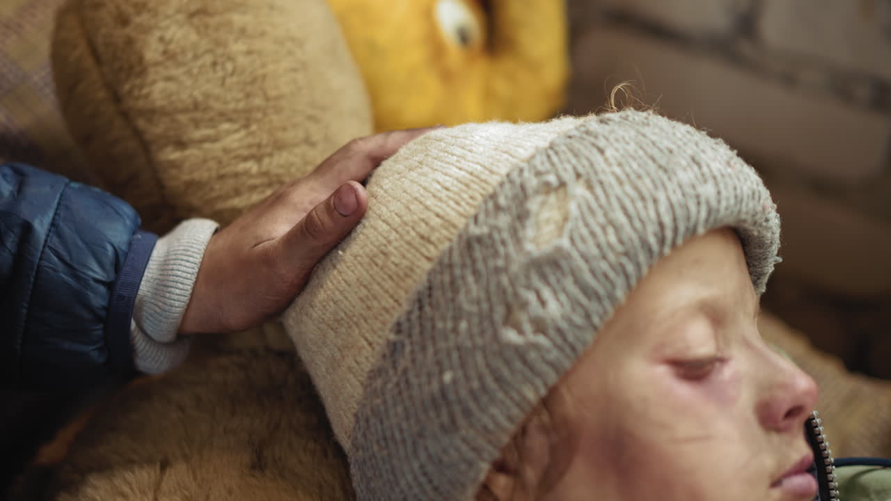 Close up of person gently tapping head of sick boy wearing worn wool hat, showing moment of care and emotional connection in harsh conditions with soft light falling over bruised face