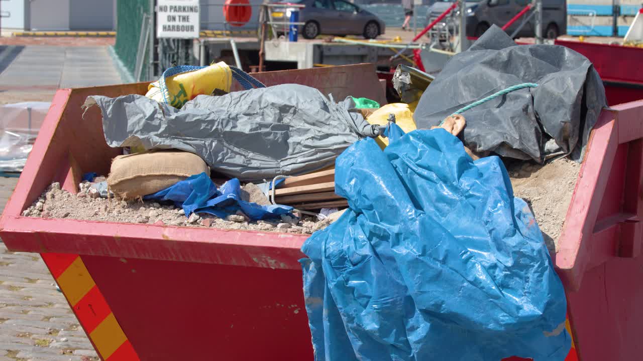Blue plastic tarp flaps in wind atop construction debris skip, daylight, static camera, urban street