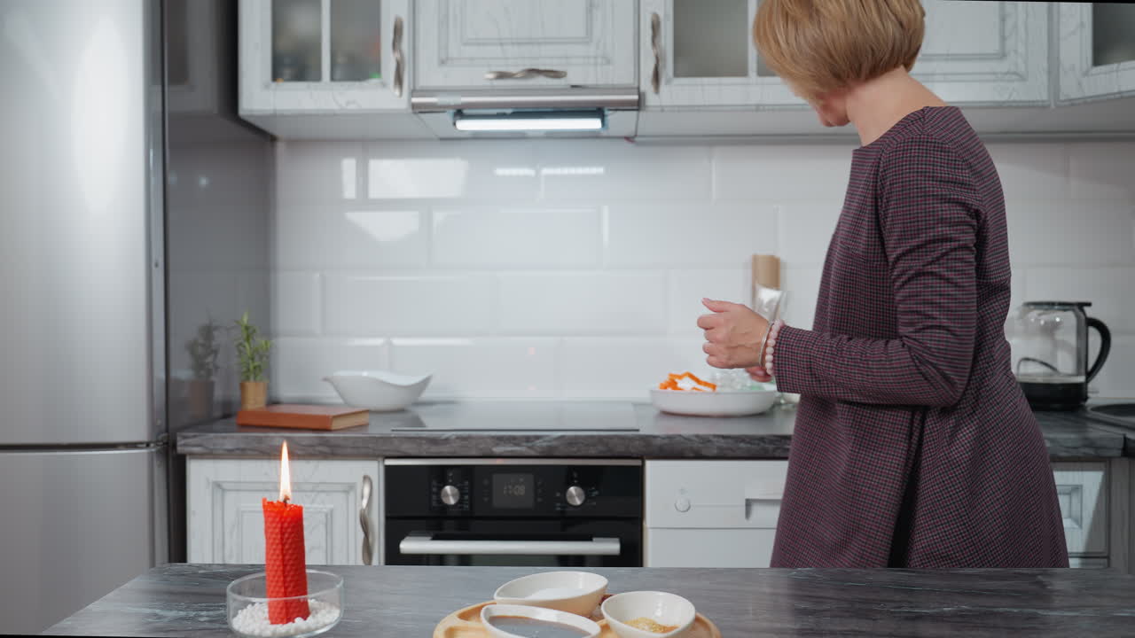 Bir kadın, modern mutfakta yemek masasını hazırlıyor; tezgahın üzerinde yanan kırmızı bir mum var, tabakları ve malzemeleri düzenliyor, titrek mum ışığı ve sıcak dekorla davetkar bir atmosfer yaratıyor.