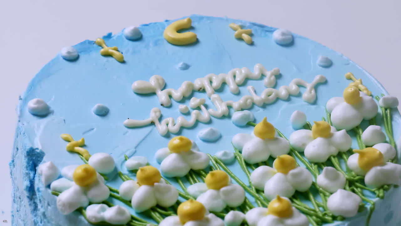 zoom de un primer plano de un pastel de cumpleaños de hielo azul, decorado con pequeñas flores blancas con puntas amarillas y acentos verdes