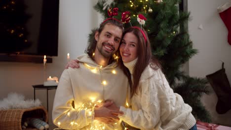 portrait of a happy couple, a brunette guy and a girl in a white sweater and in new year's hoops with a smile, holding hands and hugging each other, decorated with a new year's glowing garland near a green christmas tree in a cozy room in a winter evening