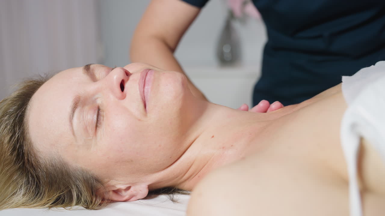 Client displays serene smile and closed eyes while skin care expert glides fingertips from side of neck to shoulder applying gentle pressure during relaxing spa session with soft ambient lighting