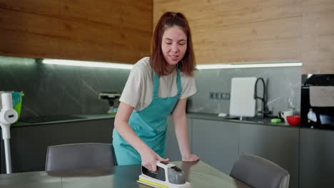 Portrait-of-a-happy-brunette-cleaning-lady-girl-in-a-blue-apron-polishing-her-table-in-the-kitchen-and-then-looking-up-and-posing-in-a-modern-kitchen-while-doing-on-call-cleaning-in-an-apartment