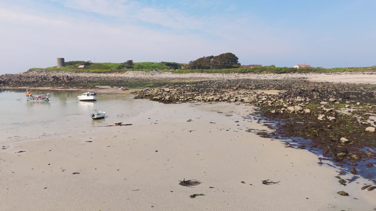 Low reveal drone footage of small boats drying out on beautiful golden sandy beach in Guernsey on bright sunny day with headland and tower in the background