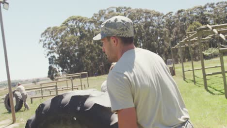 soldado caucásico en una gorra rodando un neumático de tractor en una carrera de obstáculos al sol