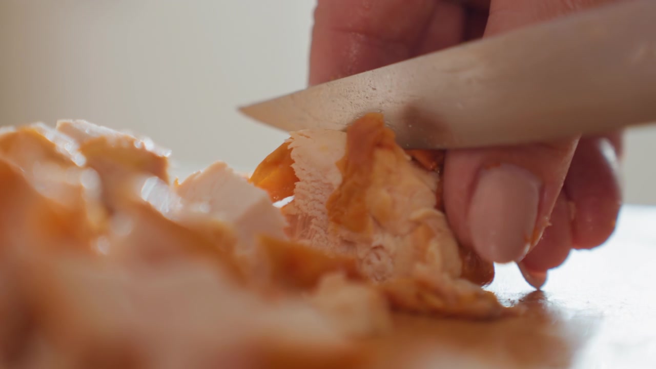 Närbild som visar en hand som styr en kniv för att skiva kokt kyckling på en skärbräda av trä, med synlig nedåtgående skärrörelse som producerar små hackade bitar och färska juicer i en varm inomhusmiljö.