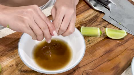 hands squeeze lime wedges into a white bowl of sauce on a wooden cutting board, with kitchen knives visible. bright, natural lighting, steady overhead shot