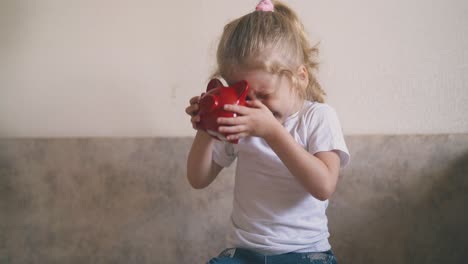 little girl in white looks into red piggybank hole on top