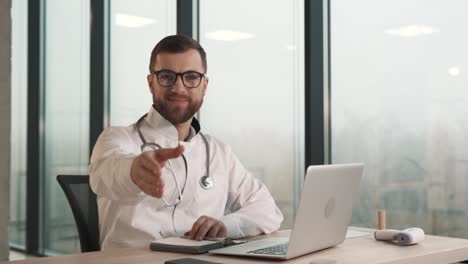 un médico masculino sólido, sentado en una mesa con una computadora portátil, le da la mano a un cliente virtual