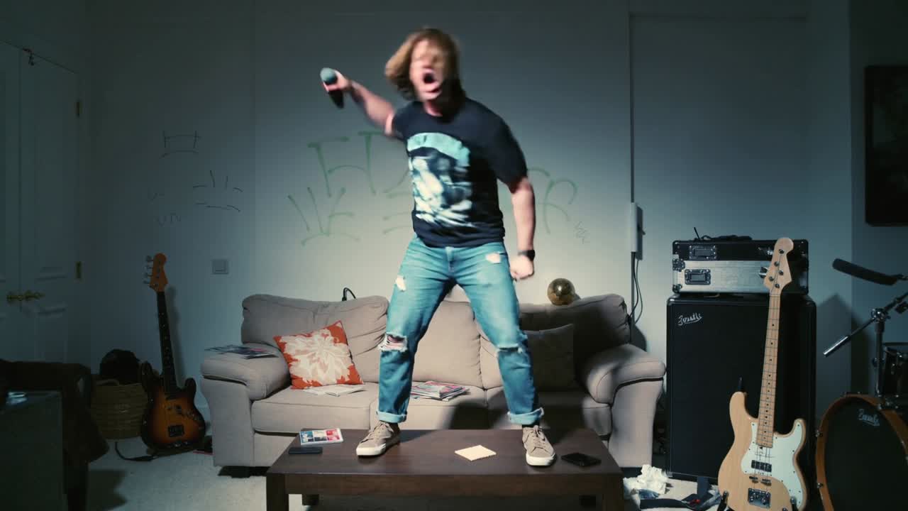 Climbing onto coffee table, man rehearsing solo in living room, gripping mic, wearing black tee