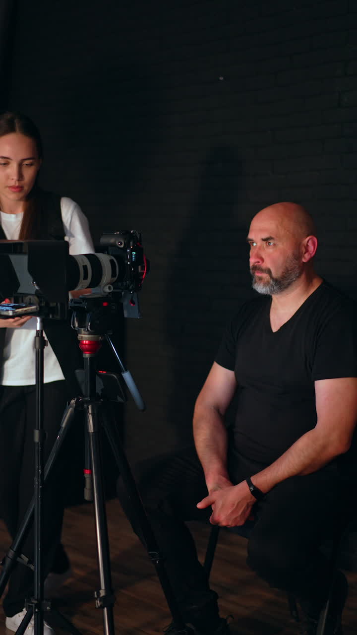 Mid-aged man wearing black t-shirt sits in studio looking at camera. Woman comes up and starts to take video on the phone. Footage backstage. Vertical video.