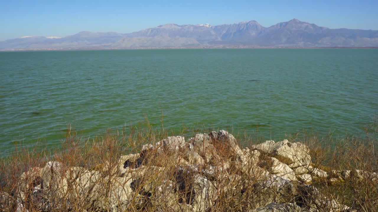 Vibrant water of lake seen from rocks and dry bushes of coast with high mountains background on a sunny winter day
