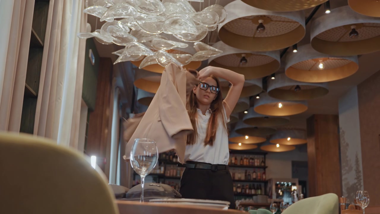 Mujer esperando en una mesa de restaurante, gafas en primer plano, mirando hacia la ventana, ajustando la postura, suave iluminación de araña, estantes con botellas y libros, ambiente relajado e introspectivo, lujo sutil