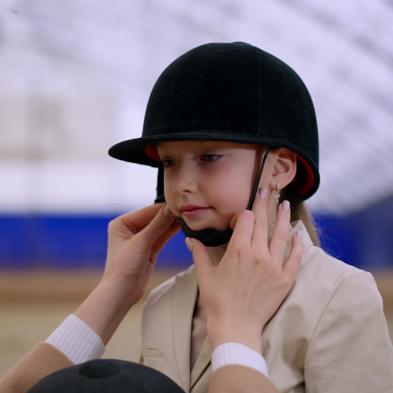 Loving mom puts carefully the jockey helmet on her daughter's head. Lovely girl prepares to horse riding at drill-hall. Close up. Blurred backdrop