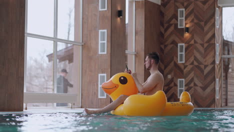 fun and rest in water park adult man is sitting on inflatable duck in swimming pool and using smartphone