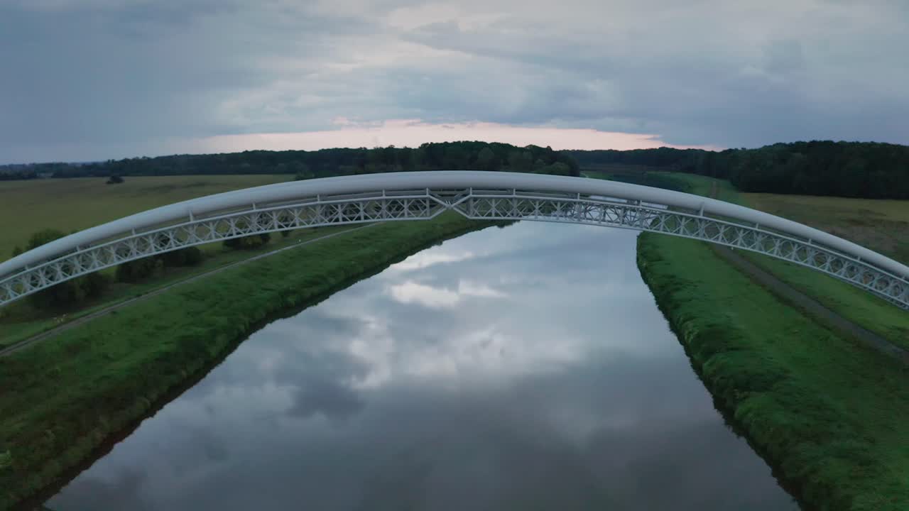 Flying above and below gas pipes across calm river Morava. Aerial sunset footage of two parallel bridges connecting both river banks.