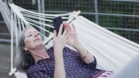 caucasian middle aged woman using smartphone relaxing in hammock on terrace in nature, slow motion