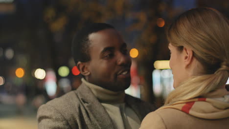 rear view of young caucasian woman hugging a african american smiling man in the street in the evening