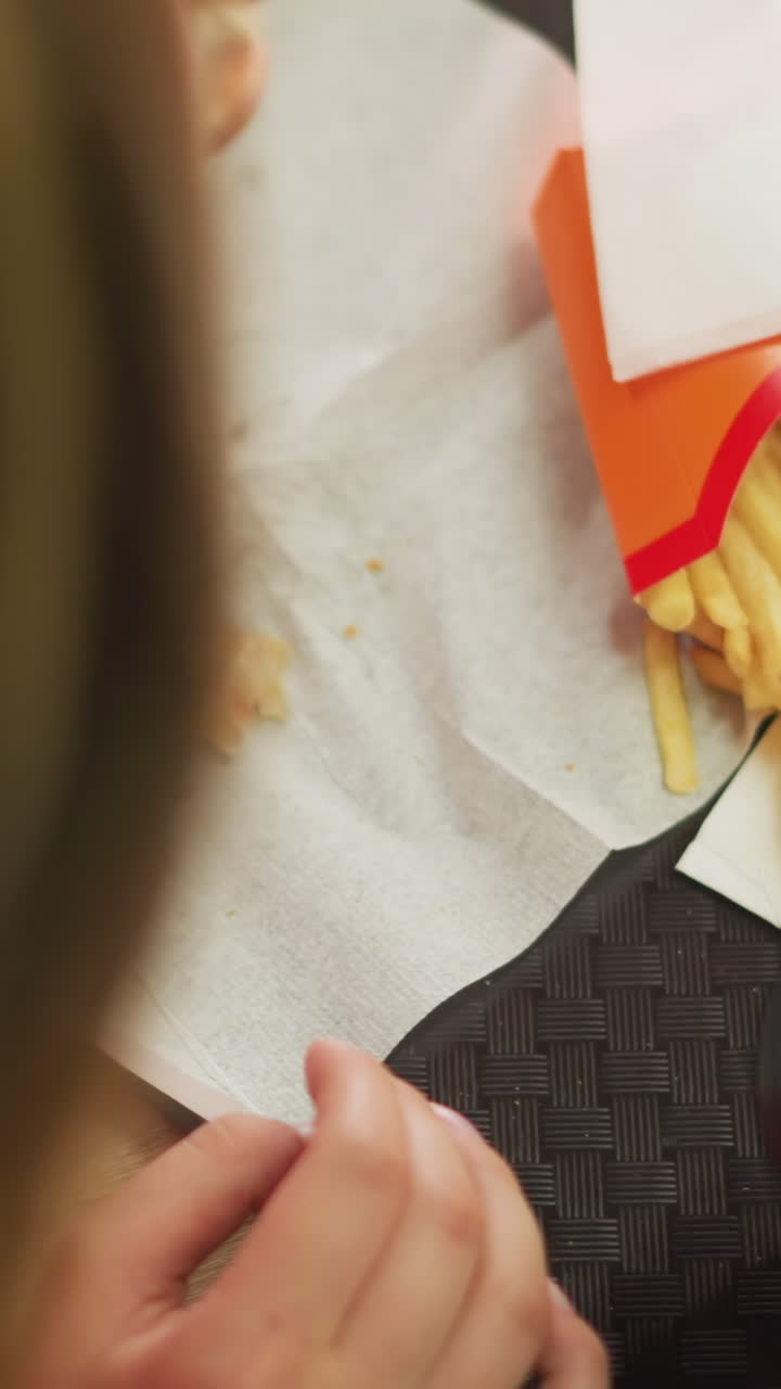la mano de la mujer recoge suavemente una hamburguesa de una servilleta colocada en una bandeja, ya que las piezas de la hamburguesa han caído en la servilleta, la bandeja también contiene papas fritas y una taza de café colocada en la mesa de madera