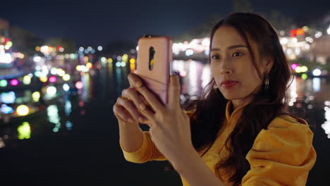 Woman-in-yellow-Ao-Dai-taking-selfie-with-lit-boats-in-Hoi-An-at-night,-blurred-background