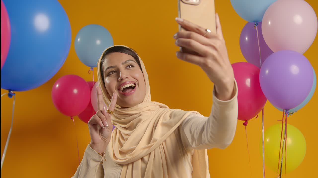Studio Portrait Of Woman Taking Selfie Wearing Hijab Celebrating Birthday Party Surrounded By Balloons