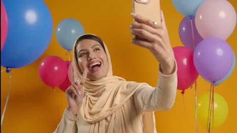 Studioporträt-Einer-Frau,-Die-Ein-Selfie-Mit-Hijab-Macht-Und-Eine-Geburtstagsfeier-Feiert,-Umgeben-Von-Luftballons