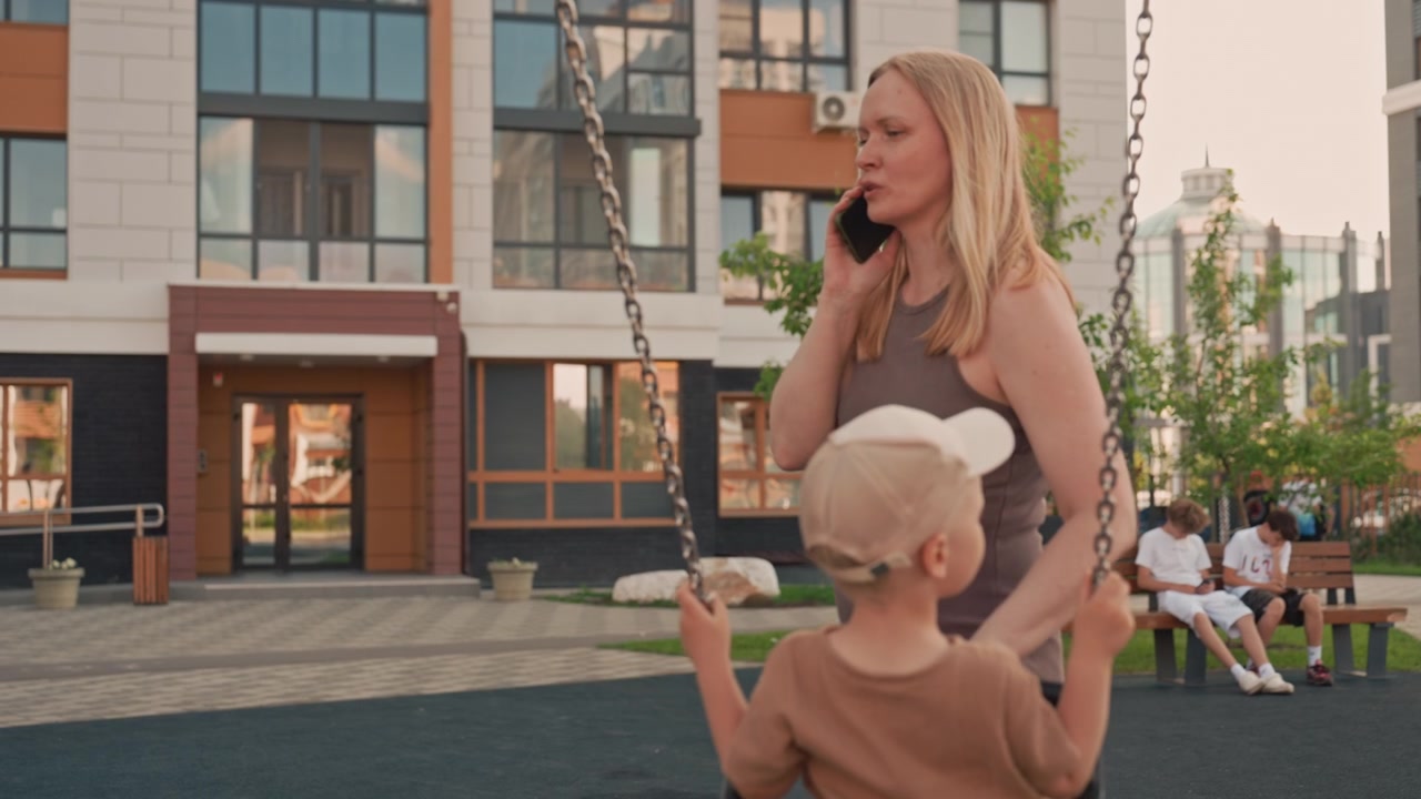 Woman talks on her phone while gently pushing young boy in swing at outdoor playground. scene takes place near modern apartment buildings with other children sitting nearby capturing