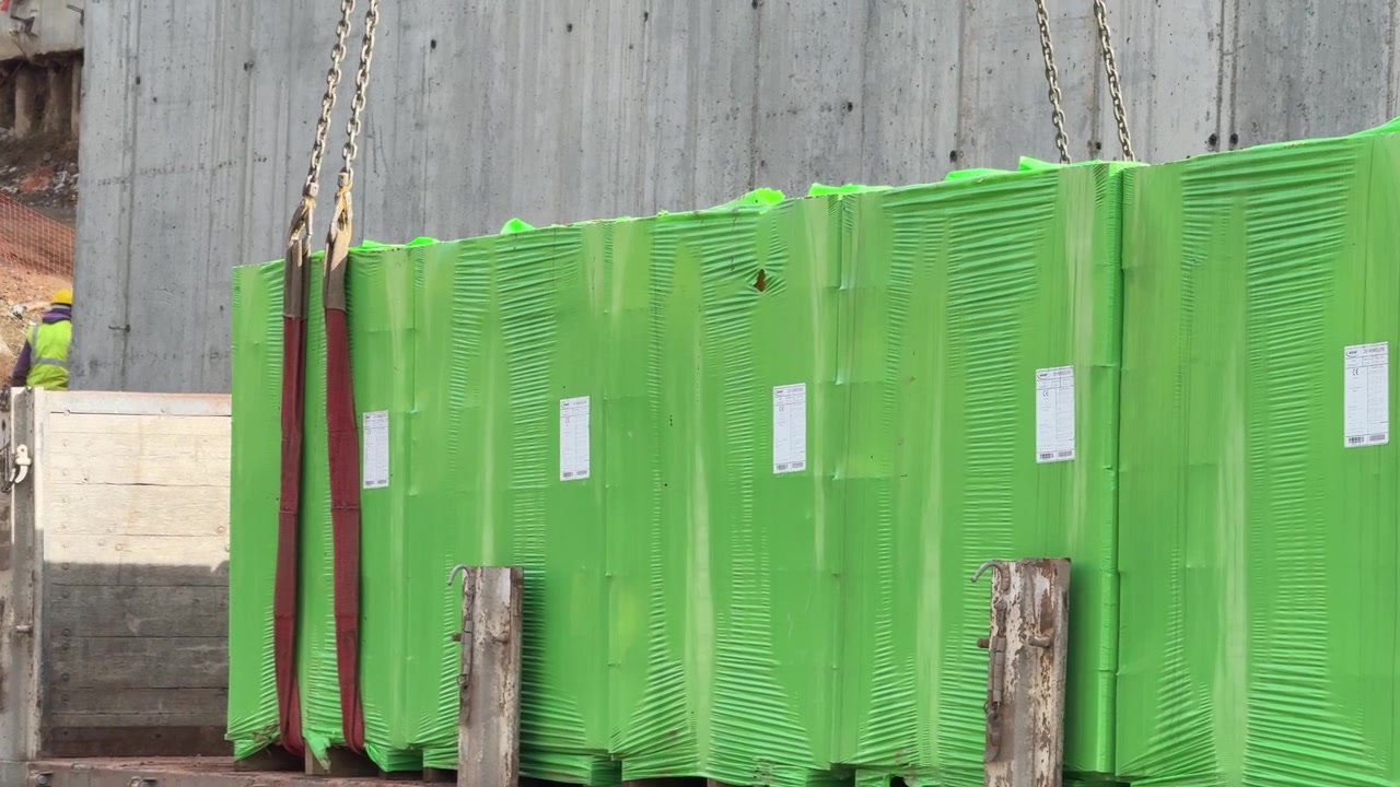 Blocchi di cemento cellulare avvolti in verde vengono sollevati dal pianale di un camion con catene e imbracature di una gru in cantiere [cite: AAA_3.jpg]. Logistica e catena di approvvigionamento dei materiali da costruzione.