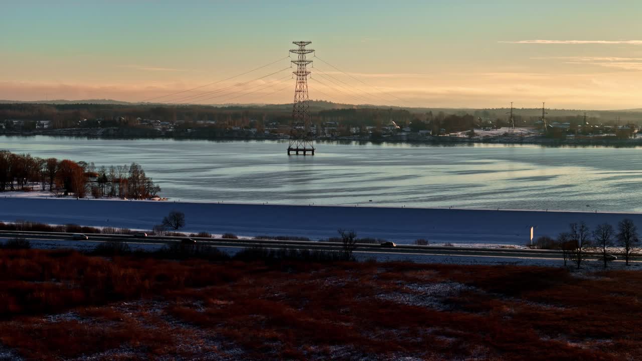 Paesaggio invernale di un fiume parzialmente ghiacciato con un traliccio di trasmissione e una città distante sotto un caldo cielo al tramonto