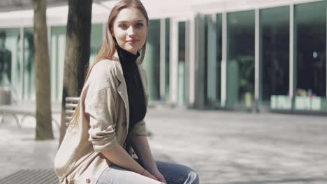 Smiling-beautiful-female-in-coat-sitting-on-bench-and-looking-at-camera