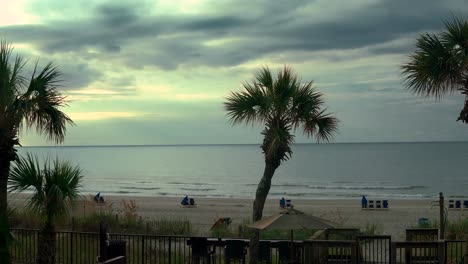 Waving-ocean-in-the-distance-with-palm-trees-on-a-cloudy-afternoon-and-beach-chairs-in-the-sand