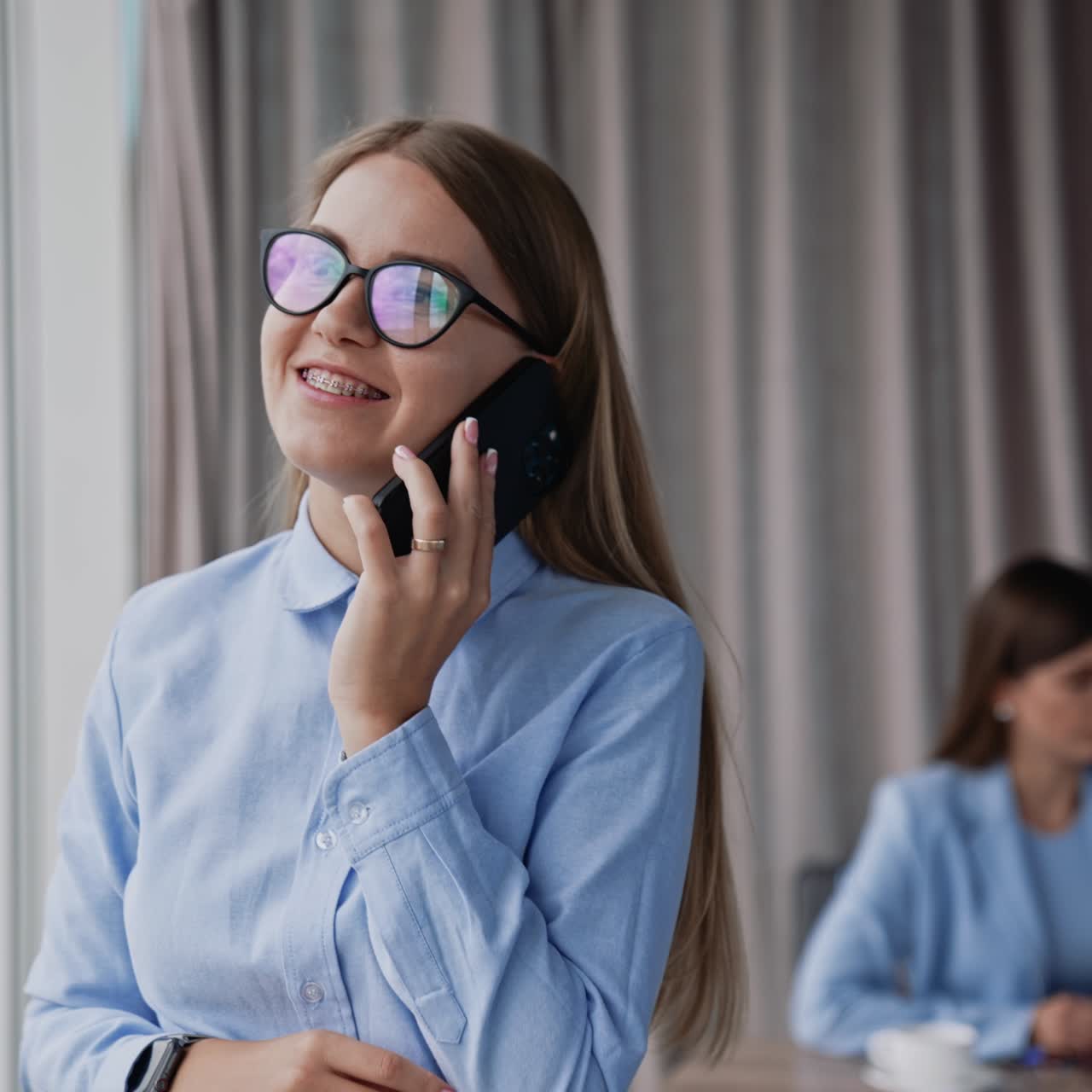 Smart young female in glasses and blue shirt starting phone conversation coming up to the window. Colleagues sitting at desk at backdrop