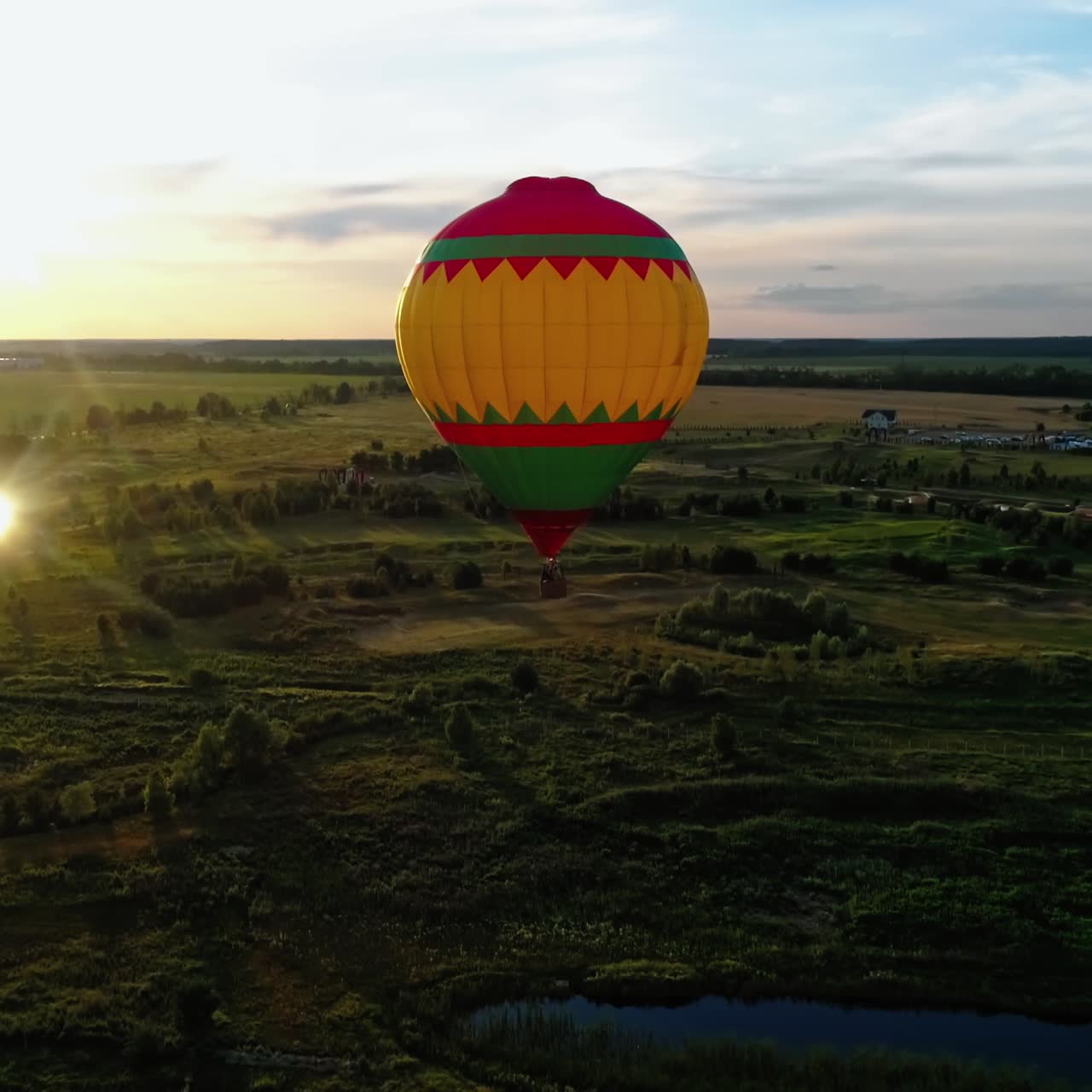 Beautiful aerostat against the evening sunlight. Colorful round hot air balloon flying over the green nature. Airship in the air in rural place at sunset. Aerial view.