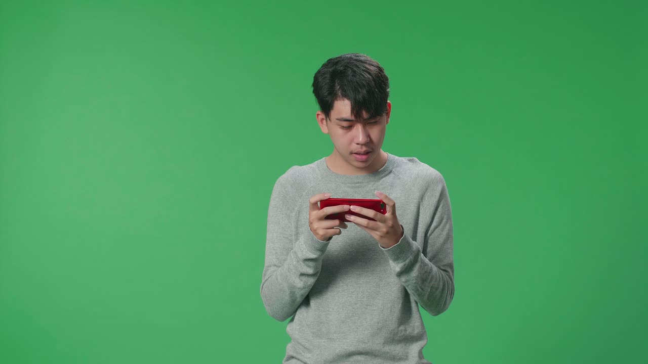 A Smiling Asian Man Using Mobile Phone Playing Game While Standing In The Green Screen Studio