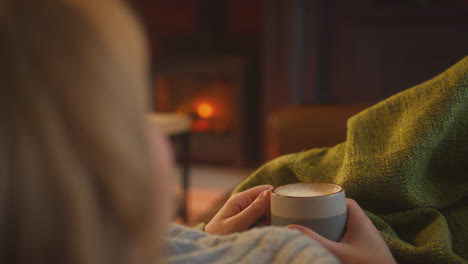 소파 에 누워 있는 라운지 에서 집 에 있는 여자 가 뜨거운 음료 를 마시며 편안 한 불 을 보고 있다