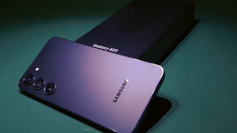 business smartphone in a studio close up that is samsung galaxy s23 black phone with three cameras in the back. placed on a dark green background while the phone leans against the box. unboxing phone.