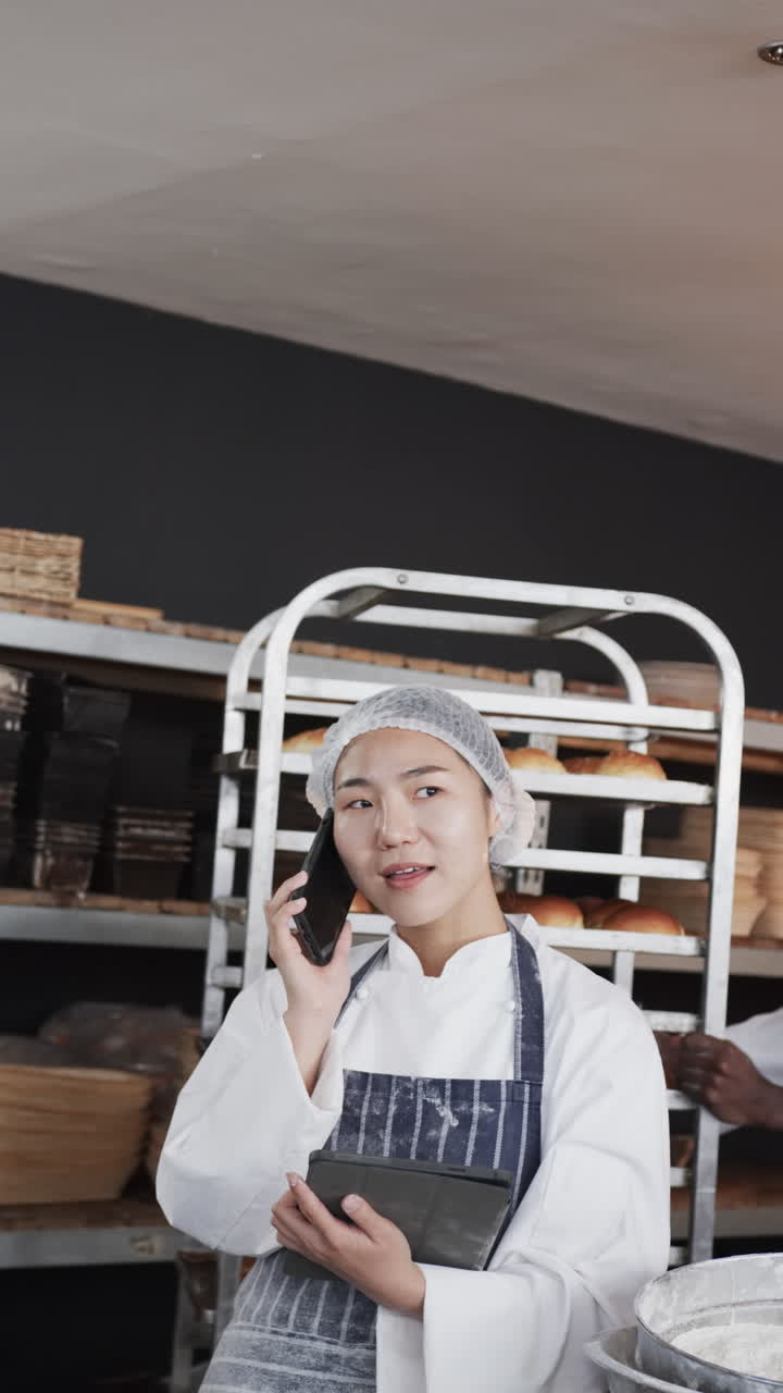 Video vertikal pembuat roti wanita Asia berbicara di ponsel pintar di dapur toko roti, gerak lambat