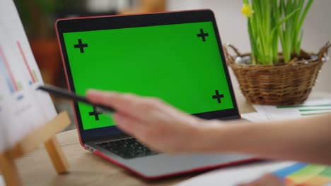 close-up laptop with green screen on table with female caucasian hand pointing away gesturing with pen. unrecognizable woman conferencing online indoors on covid-19 pandemic. freelance and remote work.