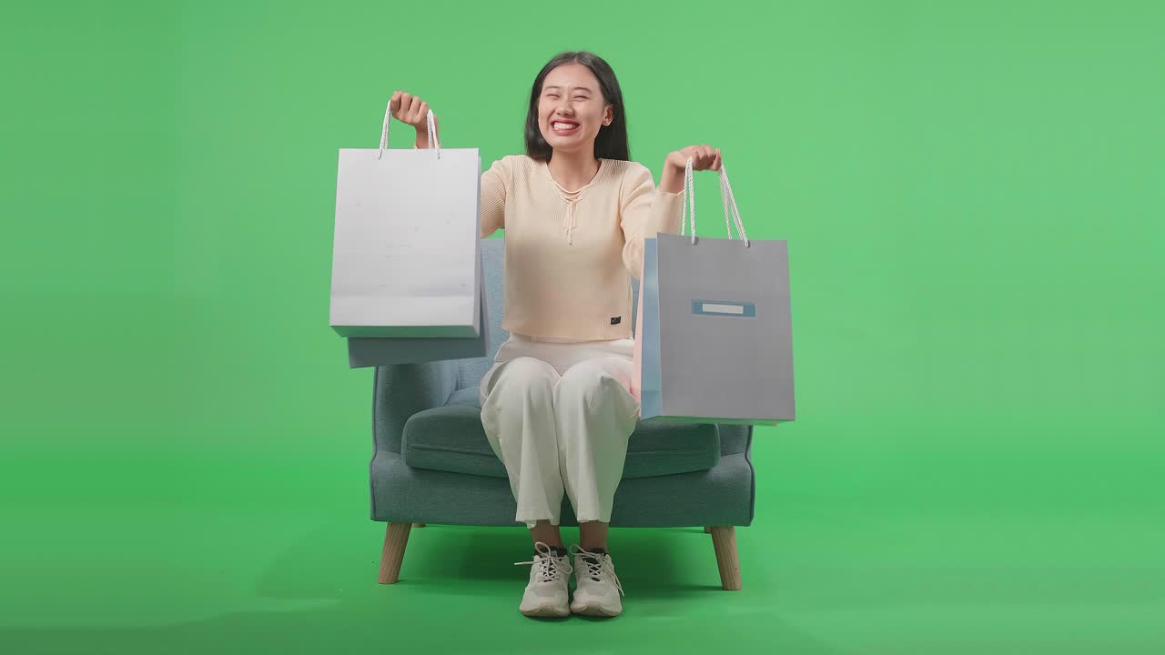 Front View Of An Asian Shopping Woman Holding Shopping Bags Up Smile To Camera While Sitting On Sofa In Front Of Green Screen