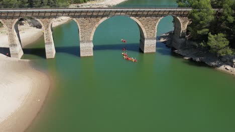 group of kayaks passing under a railway bridge over green waters