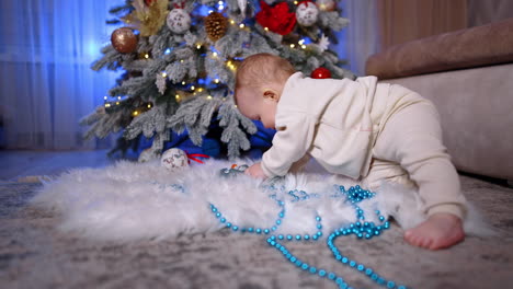 barefoot baby in white suit crawls to the soft fluffy mat under the christmas tree. toddler picks a toy and pulls it to his mouth.