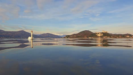 Unique-low-angle-view-from-water-surface-of-beautiful-white-swan-swimming-towards-camera-with-fortress-in-background