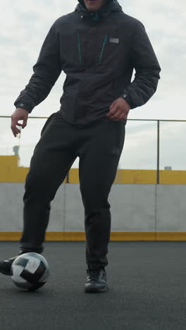 hombre controlando hábilmente la pelota de fútbol con los pies en la arena deportiva durante la sesión de entrenamiento, el fondo presenta edificios urbanos, esgrima y postes de portería