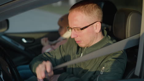 close-up of a man in a green jacket sitting in the driver s seat of a car as he fastens his seat belt. in the background, a young girl wearing a pink knit hat and white scarf is also seated in the car