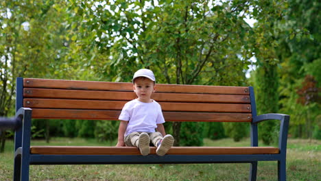 little baby boy wearing white t-shirt and cap sits on the bench. adorable child resting in the green park in summer.