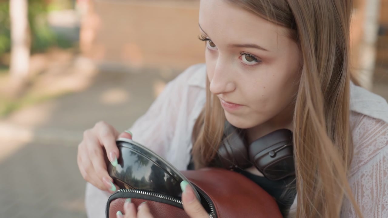 Primo piano di una giovane donna che tiene una borsa marrone con le cuffie al collo, mentre apre la cerniera di un contenitore all'interno, in un'ambientazione esterna sfocata, con un primo piano della sua camicetta bianca.