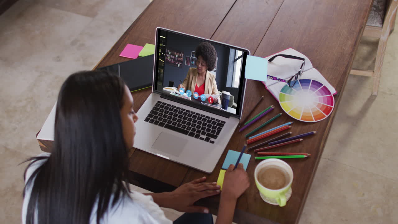 Mujer caucásica usando una laptop en videollamada con una colega, tomando notas