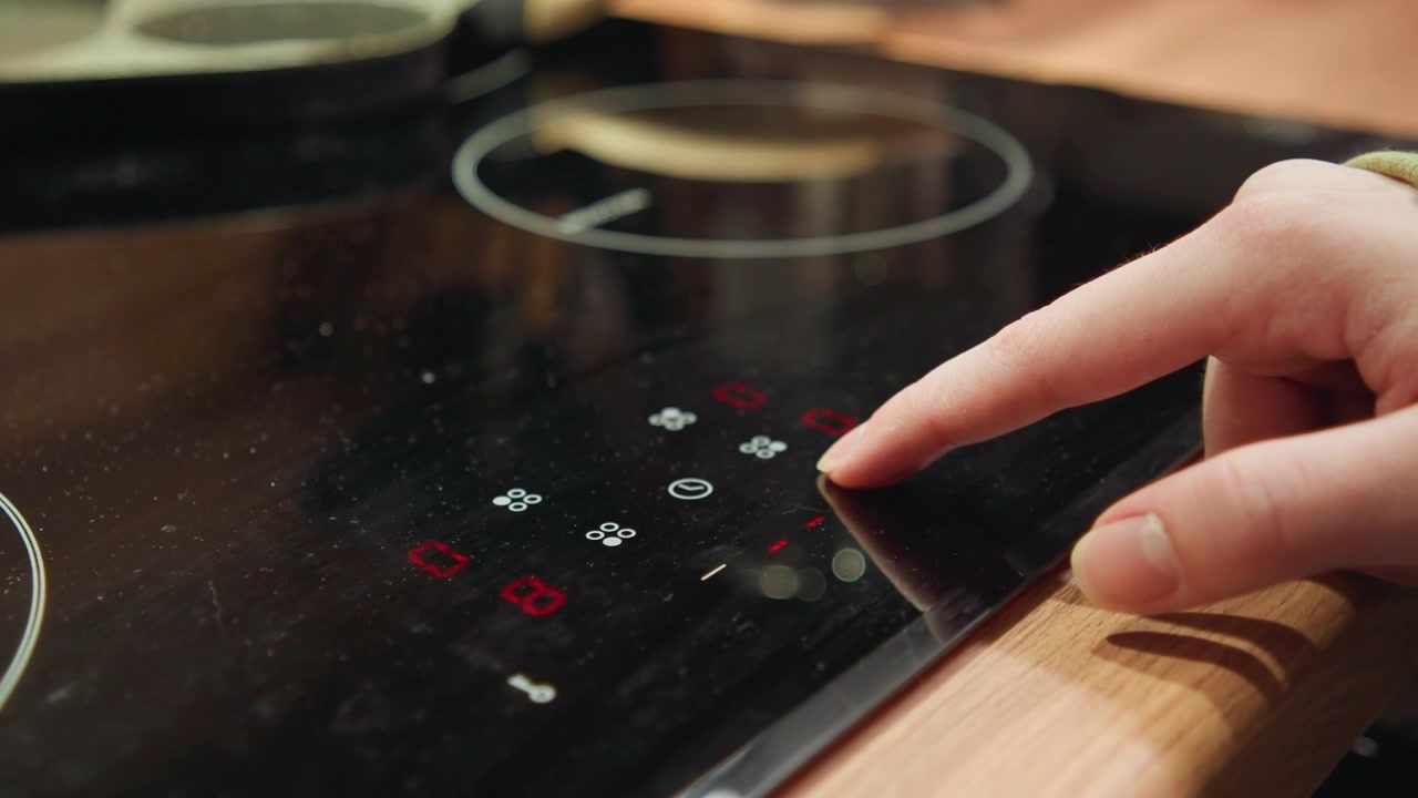 Adult hand presses illuminated touch control on glossy black induction cooktop, with glowing red and white icons, reflective ceramic surface, and modern kitchen setting suggesting cooking adjustment