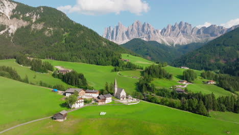 Church of Saint Mary Magdalene in countryside meadow, Val di Funes village, Dolomites valley, Odle group mountains, Drone shot, Panoramic view